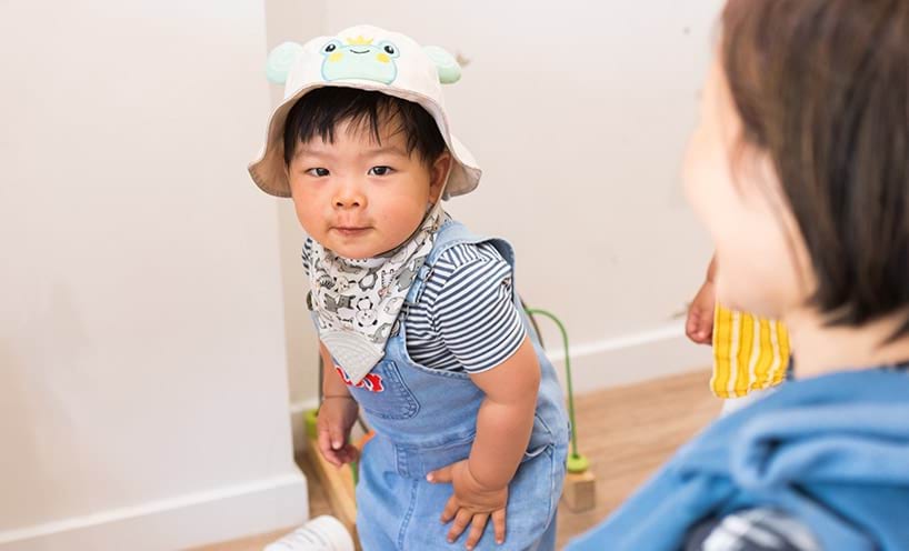 A child in a frog hat, blue overalls, a striped t-shirt and a printed neck scarf.