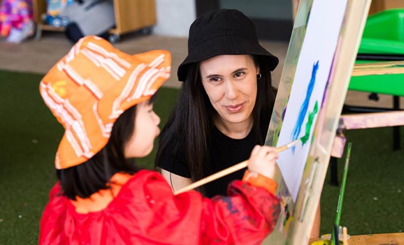 A child painting while while an educator is perched next to her.