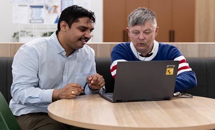 Two men sitting at a round table working on a laptop.