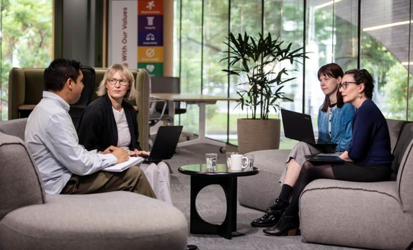 Four colleagues sitting on couches in an office with their laptops and notebooks.