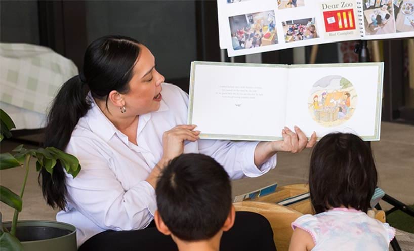A teacher sitting on the floor reading a book to 2 small children.