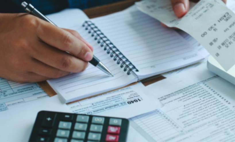 A person at a desk writing in a notepad with a calculator and paperwork work.