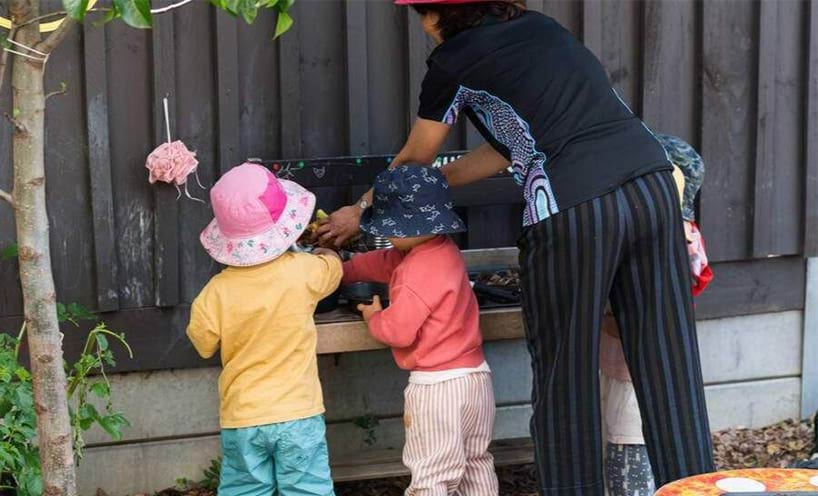 A teacher and 3 small children play at an outdoor mud kitchen.