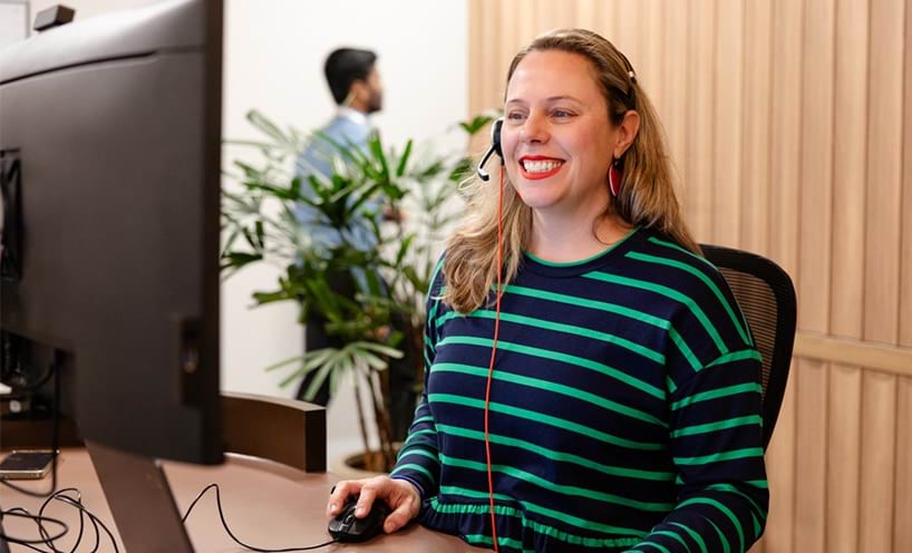 Smiling woman with a laptop, monitor and headset at a work desk in a professional setting
